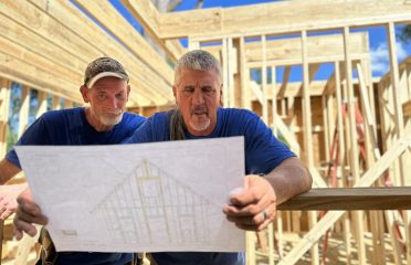 Two builders with blue shirts studying tiny home blueprint with frame in the background